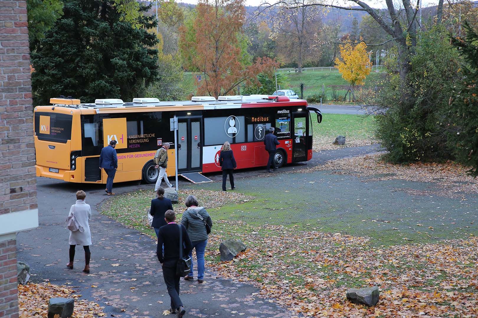 Patienten laufen auf den Medibus zu, der auf einem Parkplatz in einer Herbstlandschaft stehet.